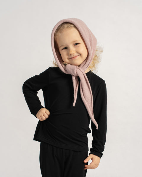Child wearing a pink knit headscarf tied under the chin, smiling and posing in a black outfit against a plain background.