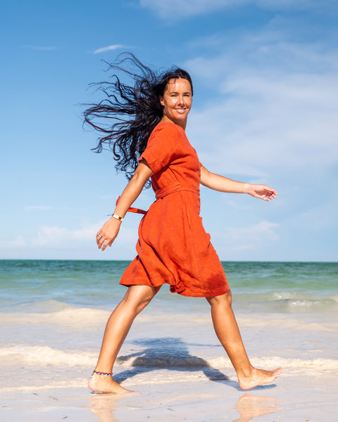 A woman with long dark hair smiles at the camera while standing barefoot on a sandy beach, holding a wide-brimmed hat above her head with both hands. She is wearing a cinnamon red wrap dress with short sleeves. The ocean is in the background with gentle waves, and several boats are visible in the distance under a bright sky.