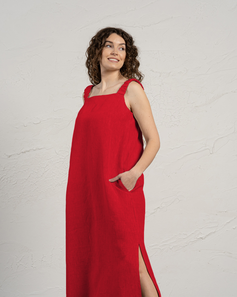 En kvinde med krøllet brunt hår står foran en struktureret offwhite væg. Hun har en ærmeløs, gulvlængde hørkjole i ren rød med brede stropper og firkantet udskæring på. Den ene hånd er stukket ned i kjolens sidelomme, og der ses et slids nederst på kjolen i den ene side. Hun kigger mod øverste venstre hjørne af billedet med et smil. Lyset er jævnt og kaster bløde skygger.