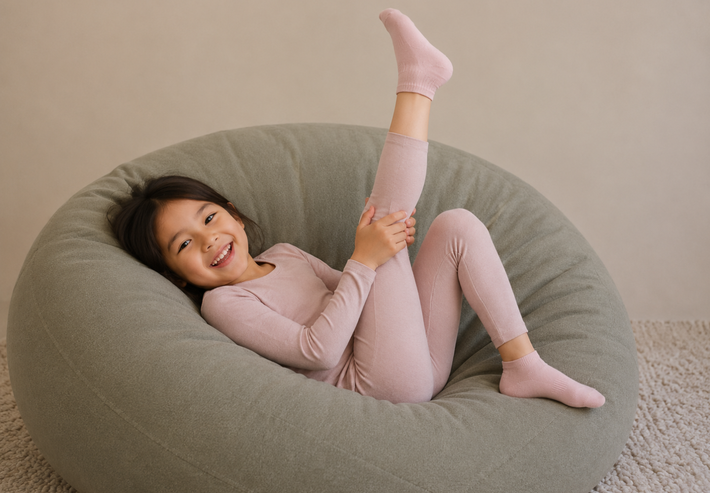 A joyful young girl relaxes in a large olive-green beanbag chair while wearing a light pink long-sleeve thermal top and leggings. She has one leg raised playfully in the air as she pulls on a matching pink ankle sock.