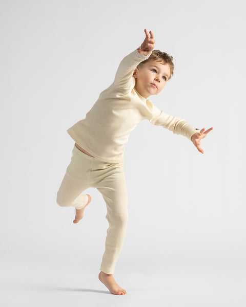 Action shot of a young boy balancing playfully on one foot, dressed in a matching cream thermal loungewear set.