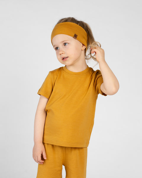 A studio portrait of a young girl standing and looking off to her left while playfully touching her hair, against a white background. She is wearing a matching set of a mustard yellow headband, short-sleeved t-shirt, and shorts.