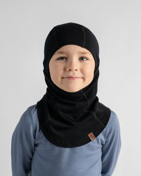 Front portrait of a smiling boy wearing a black knit balaclava covering the head and neck, paired with a dusty blue long-sleeve shirt