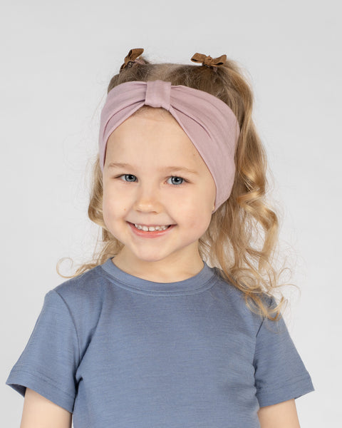 A close-up studio portrait of a young girl smiling directly at the camera. She is wearing a short-sleeved blue top and a dusty pink ribbon headband in her hair, against a plain light grey background.