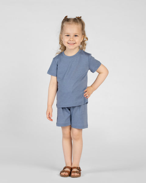 A young child wearing a dusty blue short-sleeve t-shirt, matching shorts, and brown strappy sandals smiles at the camera. They stand against a light gray background with one hand resting on their hip and their hair styled in half-up pigtails.