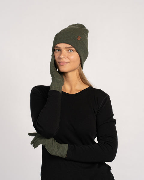 Woman wearing a dark green knit beanie and matching gloves, dressed in a black long-sleeve top, posing with one hand touching her face and the other resting across her body against a plain background.