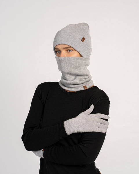 Kvinde iført en lysegrå strikket hue, matchende handsker og en halsedisse, der dækker hendes ansigt, iført en sort bluse med lange ærmer, poserer med armene over kors foran en ensfarvet baggrund.