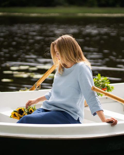 A woman in a light blue linen top and dark blue pants sits in a rowboat, gazing down softly at the sunflowers resting in her lap. One hand lightly grips the edge of the boat as she floats on the dark water.