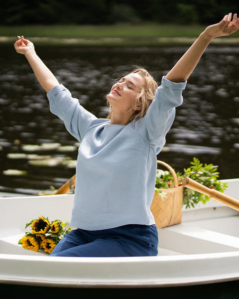 A woman sits in a white boat with her arms stretched joyfully up towards the sky, her eyes closed in relaxation. She is wearing a light blue linen shirt and dark blue pants, accompanied by bright sunflowers on the seat next to her.