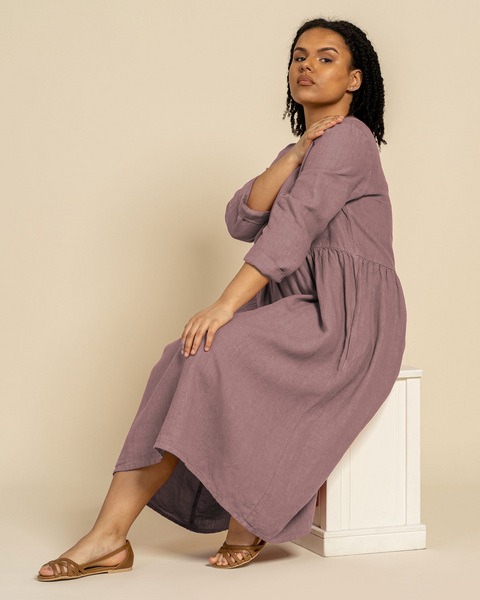 En kvinde med mørkt krøllet hår sidder på en hvid blok og ser mod kameraet. Hun har en langærmet menique hørkjole og sandaler på. Hendes venstre hånd hviler på hendes højre skulder. Baggrunden er ensfarvet lys brun.