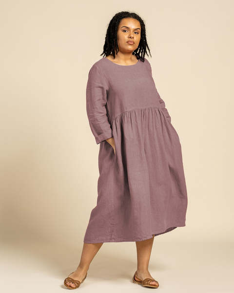 En kvinde med mørkt krøllet hår står og ser mod kameraet iført en menique hørkjole med lange ærmer i farven falmet rose. Hendes hænder er i kjolens sidelommer. Hun står foran en ensfarvet lys brun baggrund.