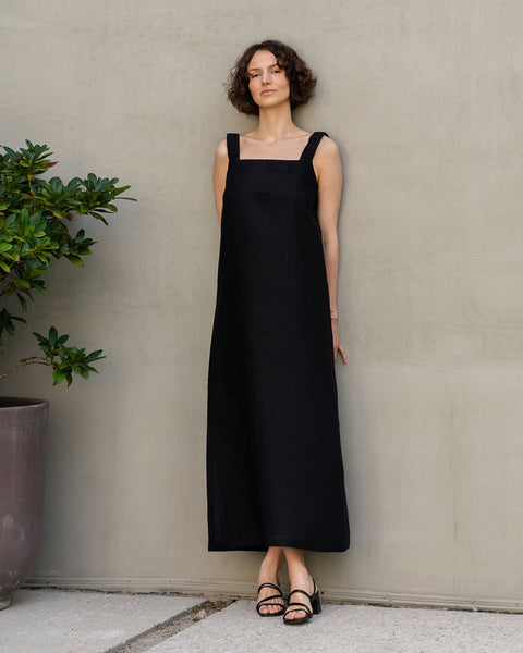 En lys hudet kvinde med kort, krøllet mørkt hår står op ad en struktureret lys brun væg. Hun har en ærmeløs, lang hørkjole med firkantet halsudskæring og sorte sandaler på. Til hendes venstre side står en stor grøn plante i en potte.