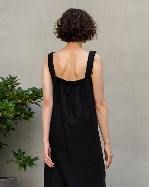 En lys hudet kvinde med kort, mørkt krøllet hår står med ryggen mod beskueren. Hun har en ærmeløs, helt sort maxikjole i hør på, med rynkede detaljer bagpå. Til hendes venstre side står en grøn plante i en potte, og hun står op ad en ensfarvet, lys væg.