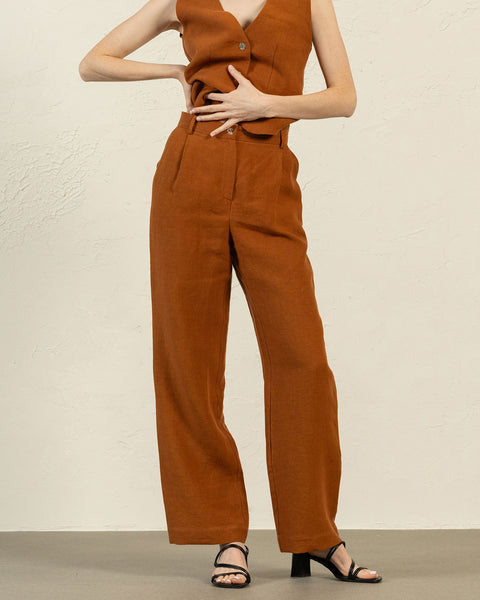 En person ses fra midt på overkroppen og ned, iført en ærmeløs vest i mandelbrun hør og bukser med vide ben. Den ene hånd hviler på hoften, mens den anden blidt rører ved maven. Personen har sorte åbne sandaler med hæl på. Baggrunden er en tekstureret lys beige væg og en lys grå gulv.