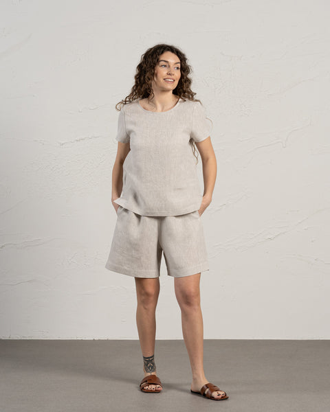 En kvinde med brunt krøllet hår iført menique 100 % hør bermudashorts og hør t-shirt sæt. Hendes hænder er i lommerne, mens hun kigger til siden. Baggrunden i billedet er lys og i kølige toner.