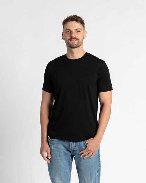 En mand med kort, mørkebrunt hår og et veltrimmet skæg står midt på en ensfarvet hvid baggrund og ser direkte mod beskueren. Han bærer en sort t-shirt med rund hals og korte ærmer fra menique-mærket, der fremhæver tøjets pasform og enkelhed. T-shirten er lavet af merinouldstof. Han har også lyseblå denimbukser med lige ben og en synlig forlommer, hvor den ene hånd afslappet er stukket ned i lommen.