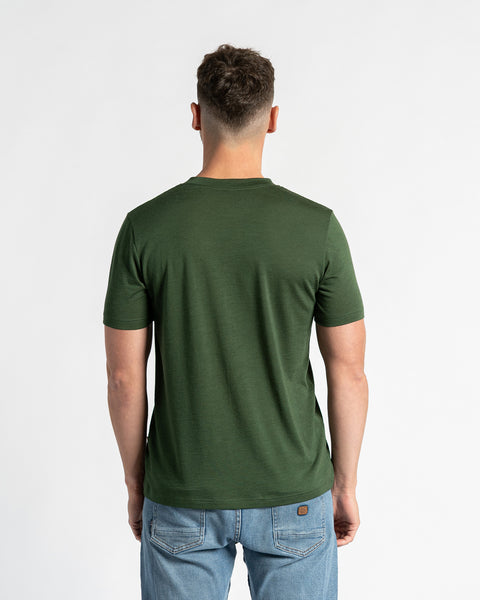 Bagfra billede af en mand i en mørkegrøn t-shirt af merinould og lyseblå cowboybukser, stående mod en hvid baggrund.