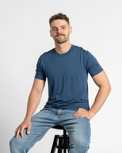 En mand iført en t-shirt i denimfarve og lyseblå jeans sidder på en skammel mod en lys baggrund. Han har kort brunt hår, et let skæg og smiler, mens hans hånd hviler på hoften. Merinould-t-shirten er i fokus og viser dens pasform og udseende, mens manden sidder ned.