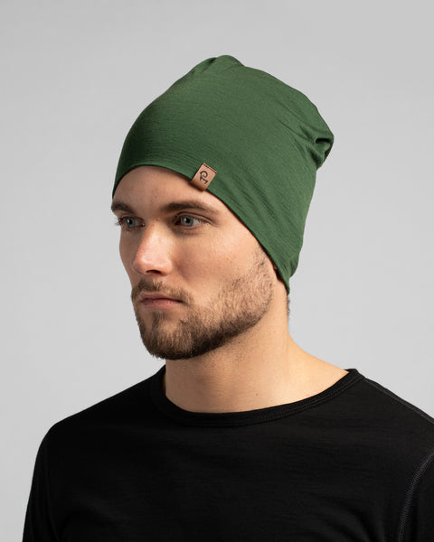 A studio portrait of a man looking off to the left against a light grey background. He is wearing a black long-sleeved shirt and a dark green slouchy beanie featuring a small brown logo tag.