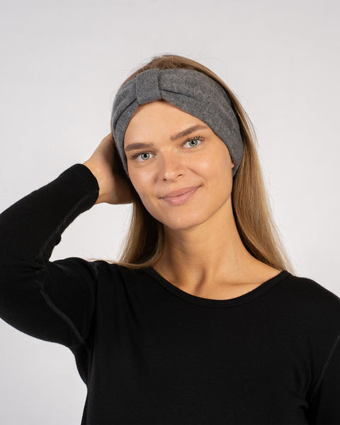 Kvinde iført et mørkegråt pandebånd med en knude foran, klædt i en sort bluse med lange ærmer, smilende blidt mens hun rører ved sit hår mod en ensartet lys baggrund.