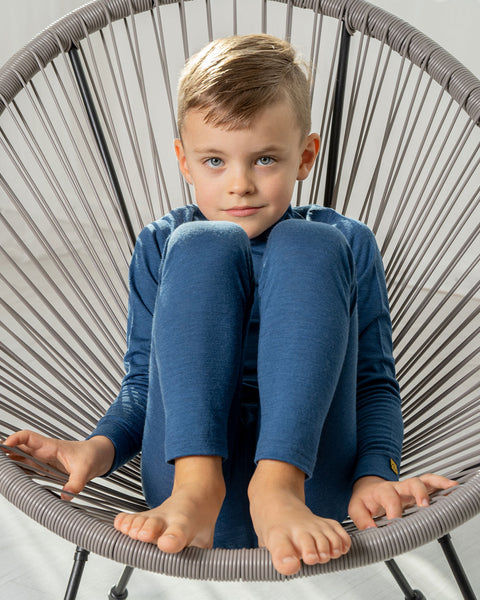 A boy sitting in a lounge chair, wearing the menique merino pants in denim color, and a matching long sleeve shirt. His look is intense, pointed at the camera. His legs are raised onto the chair, with the knees bent.