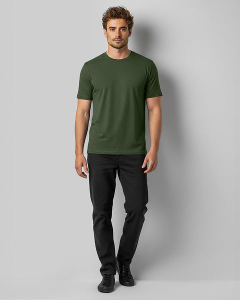 En mand står med fronten mod kameraet iført en grøn T-shirt, sorte jeans og sorte sportssko foran en ensfarvet grå baggrund.