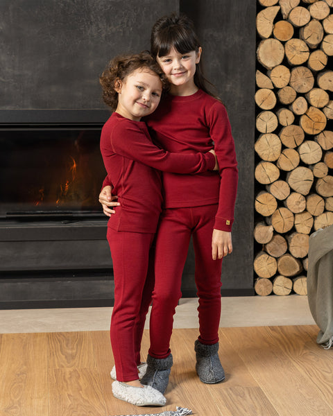 Two children, a boy and a girl, standing close to each other wearing a cherry royal merino long sleeve top and matching pants. The girl has her arms around the boy.