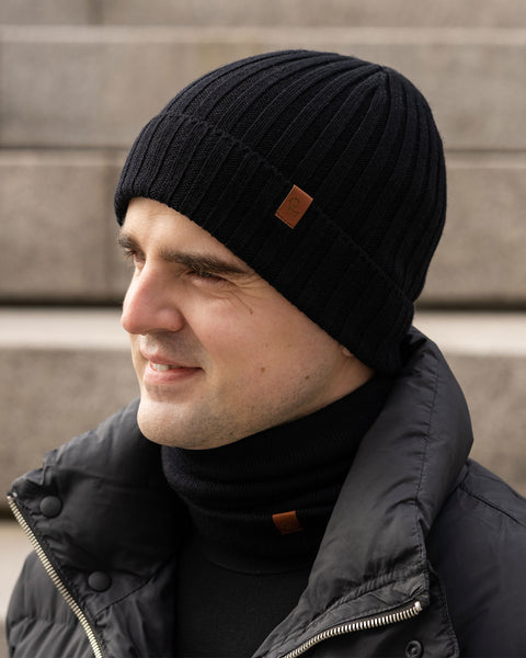 En person med lys hud vises i profil, smilende og kiggende mod venstre. De har en sort ribstrikket hue på med en lille brun mærkat, en sort rullekravetrøje, en sort strikket halsedisse med en lignende brun mærkat og en sort dunjakke. Baggrunden ser ud til at være betontrapper i et udendørs miljø.