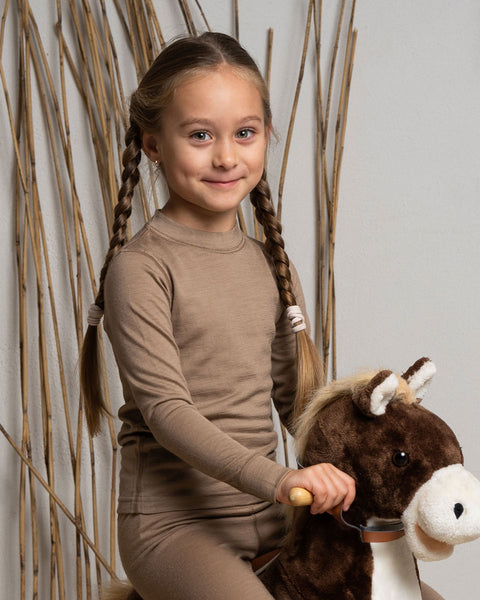 A girl wearing the menique Kids' Merino 160 Long Sleeve Crew in Beige color, riding a kids' toy horse.