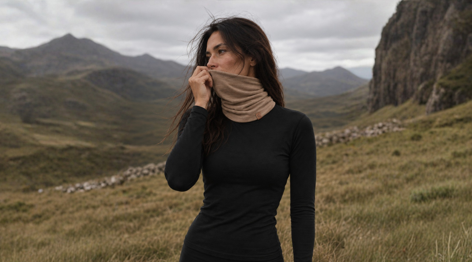 A woman with long dark hair stands in a grassy mountain landscape, pulling a tan neck gaiter up over her nose and mouth as the wind blows her hair. She is wearing a fitted black long-sleeved top under a moody, overcast sky.