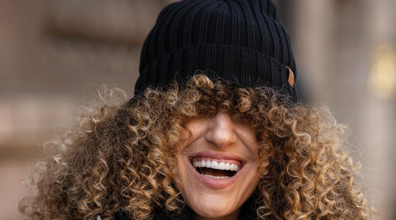 A woman wearing a black knit merino wool beanie hat, that is falling over her head, covering her eyes. She is smiling. The model has curly light brown hair.