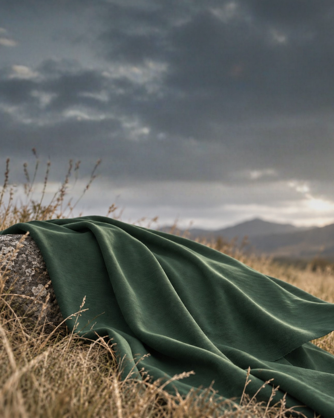 A piece of textured dark green fabric is draped over a weathered rock in a field of dry, wild grass. The fabric's edge catches a gentle breeze against a softly blurred backdrop of rolling mountains and a hazy, overcast sky.