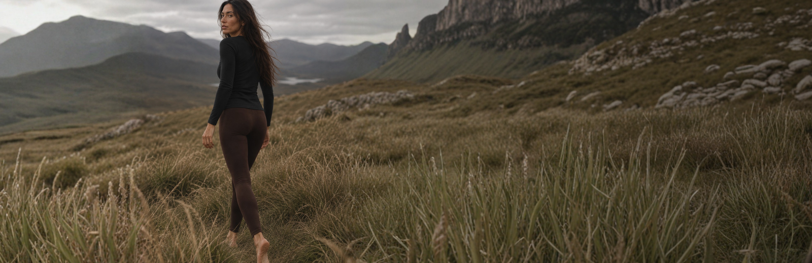 A barefoot woman with long dark hair turns to look back over her shoulder while walking through a field of tall, dry grass. She is wearing a black long-sleeved top and brown leggings, set against a mountainous landscape under a dark, cloudy sky.