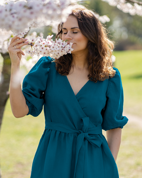 A light-skinned person with wavy brown hair has their eyes closed and appears to be smelling a cluster of white blossoms held in their right hand. They are wearing a linen menique wrap dress with short puff sleeves and a tied waist, and their left hand is in a pocket. A blossoming tree and a blurred green background suggest an outdoor setting.