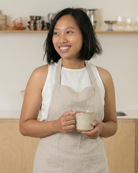 Close-up of a woman in a beige linen kitchen apron holding a coffee mug, minimalist home kitchen setting.