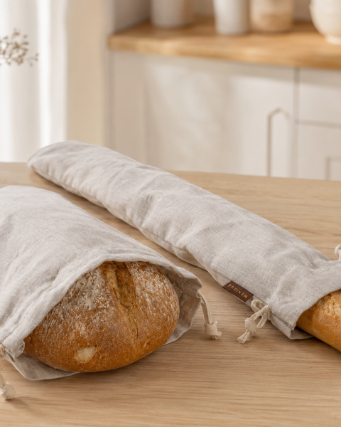 Two natural-colored linen bread bags resting on a light wooden table. A medium bag on the left holds a round artisan loaf, and a long, slender bag on the right holds a crusty baguette. Both feature cinched drawstring closures.