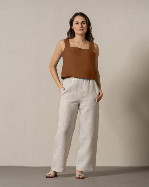 En kvinde med mellemtonet hud og mørkt, skulderlangt hår står vendt fremad mod en ensfarvet, lys væg. Hun har en tealfarvet, ærmeløs top med firkantet halsudskæring på, og linnedbukser fra Menique med den ene hånd i lommen.