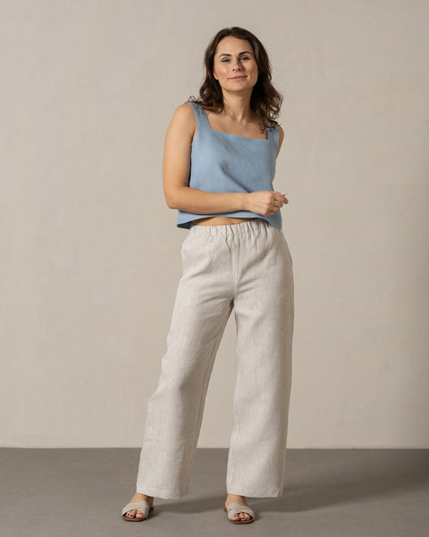 En kvinde med mellemtonet hud og mørkt, bølget hår står vendt mod kameraet med et let smil. Hun har menique hør på, en ærmeløs top med firkantet halsudskæring og hørbukser med vide ben og elastik i taljen. Hendes hænder hviler afslappet ved siden af hende, og hun har lyse sandaler på. Hun står på en lys grå overflade foran en ensfarvet, lys beige væg.