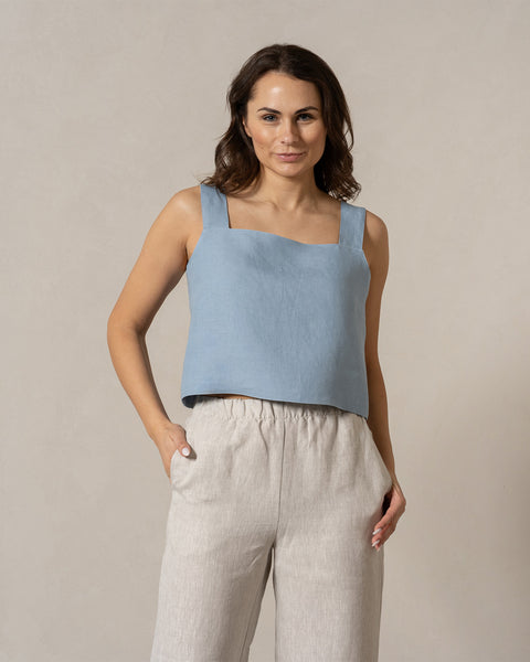 En kvinde med mellemtonet hud og mørkt, bølget hår ser direkte ind i kameraet med et blidt udtryk. Hun har en skyblå, ærmeløs top med firkantet halsudskæring på, samt naturfarvede, vidde bukser med elastik i taljen, og hendes hænder hviler afslappet i lommerne. Baggrunden er ensfarvet og lys beige.