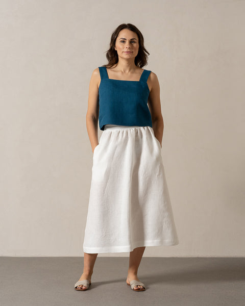 En kvinde med mellemtonet hud og mørkt, skulderlangt hår står vendt fremad mod en ensfarvet, lys væg. Hun har en blågrøn, ærmeløs top med firkantet halsudskæring på, og en hvid menique-nederdel med hænderne i lommerne. Hendes fødder er bare, og hun ser ud til at stå på et lysegråt gulv.