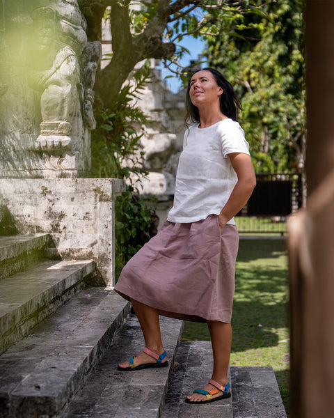 En kvinde med mørkt hår står på stentrapper udendørs og kigger opad. Hun har en hvid kortærmet bluse på, et støvet lyserødt knælængde skørt og farverige flade sandaler. Hendes hænder er i skørtets lommer. Ved siden af hende er en vejrbidt stenmur med udskæringer, og træer med grønne blade kan ses i baggrunden under en klar himmel.