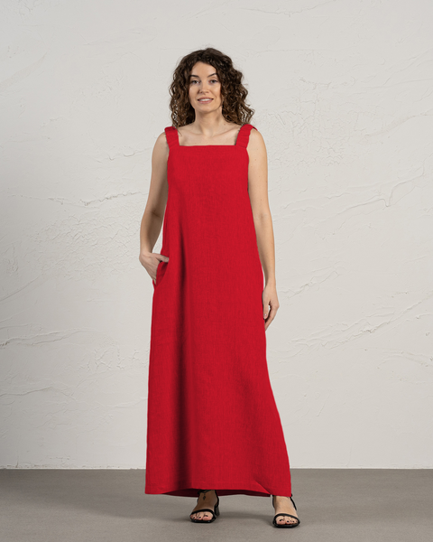 En kvinde med krøllet brunt hår står foran en struktureret off-white væg. Hun har en menique ærmeløs, gulvlængde ren rød kjole på med brede stropper og firkantet udskæring. Den ene hånd er stukket ned i kjolens sidelomme. Hun har sorte sandaler med åben tå på og kigger mod højre side af billedet med et let smil. Lyset er jævnt og kaster bløde skygger.