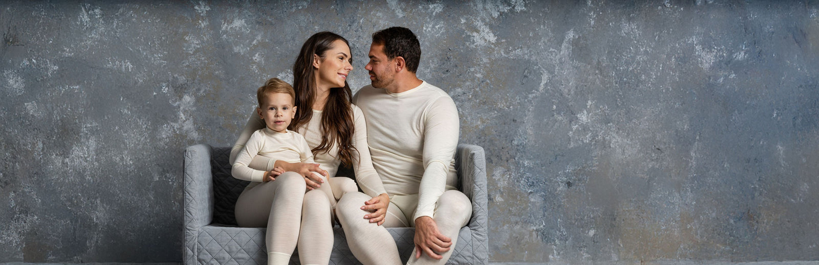 Familie på tre, der sidder sammen på en grå stol foran en struktureret grå væg.