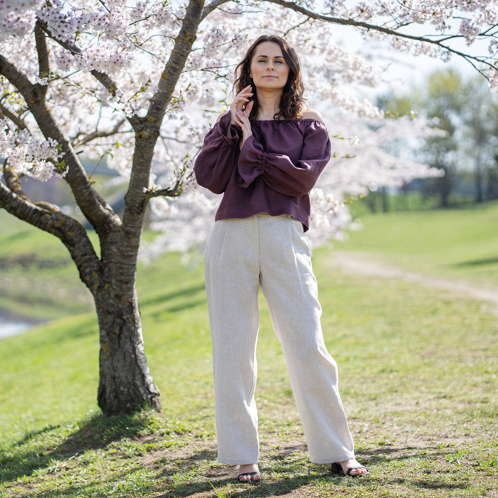 En kvinde med lys hud og mørkt hår ses fra livet og op, smilende direkte til beskueren. Hun har en skygge-lilla bluse med rund hals på. Hendes venstre hånd, prydet med en ring på ringfingeren, er afslappet løftet mod brystet, med tommelfingeren berørende hendes udskæring.