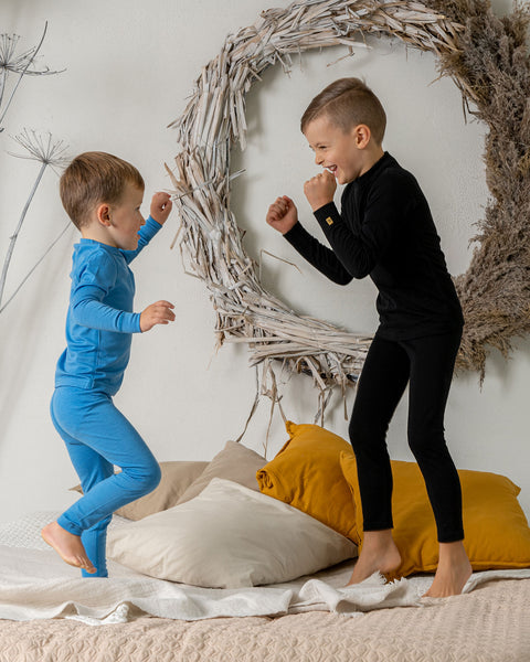 Two boys jumping on a bed, one of them is wearing the menique 160 kids' set of long sleeve  crew and pants in light blue and the other in black color. They are play-fighting.