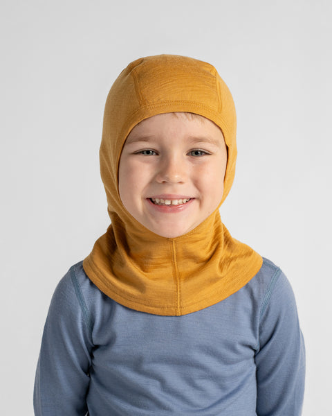 Young boy smiling at the camera wearing a bright mustard yellow balaclava paired with a light blue long-sleeve shirt.