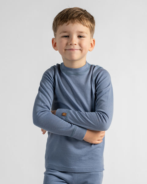 Young boy smiling with his arms crossed, wearing a long-sleeved dusty blue thermal base layer shirt against a light grey background.
