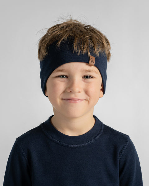 A young boy smiling directly at the camera against a light gray backdrop, wearing a navy blue t-shirt and a matching navy knit headband.