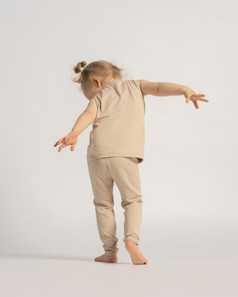 Lille pige i et beige ærmeløst sæt med top og leggings set bagfra, gående barfodet på en lys baggrund.