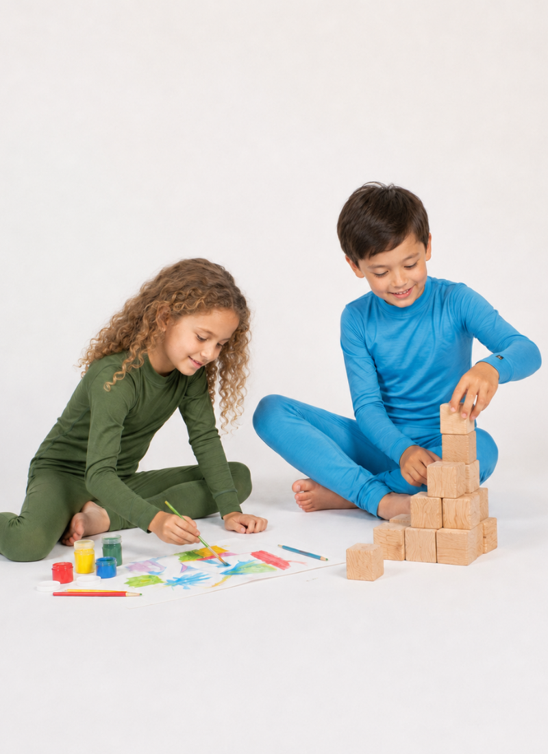 Two barefoot children sitting on a white floor playing. A girl on the left wearing an olive green long-sleeved base layer set paints a colorful picture, while a boy on the right wearing a blue long-sleeved base layer set builds a pyramid out of wooden blocks.